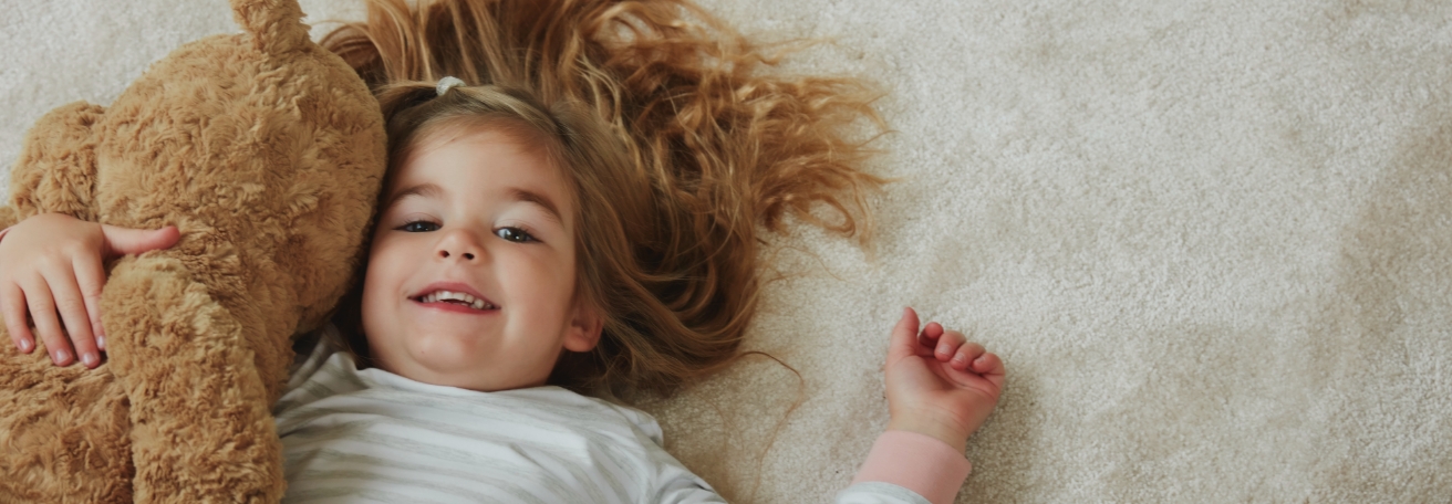 A young girl with blond hair cuddles a teddy bear while lying on a cream carpet floor